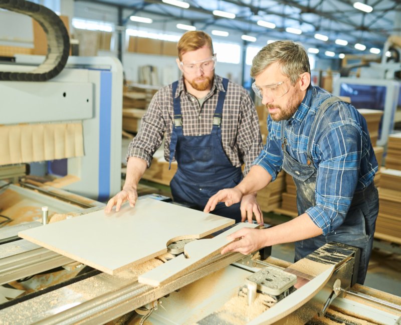 Serious concentrated factory workers in uniform standing at workbench and joining wooden pieces while assembling furniture at factory